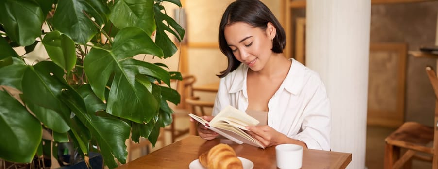 Smiling young asian woman enjoys reading book while sits alone in cafe, drinks coffee, looking at page.