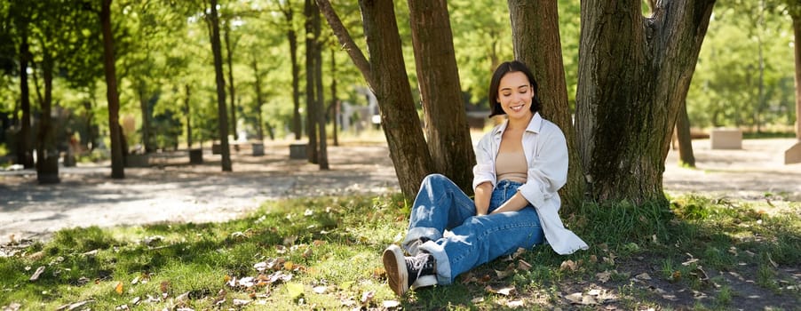 Young people. Beautiful asian girl sits near tree in park and rests, smiling and looking into distance, relaxing outdoors on fresh air in summer.