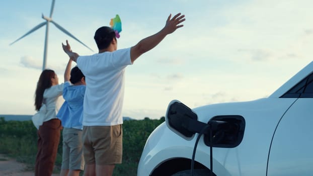 Concept of progressive happy family enjoying their time at wind farm with electric vehicle. Electric vehicle driven by clean renewable energy from wind turbine generator for charging station.