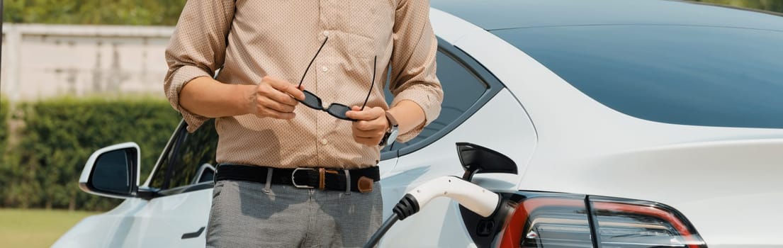 Young man recharge electric car's battery from charging station in outdoor green city park. Rechargeable EV car for sustainable environmental friendly urban travel. Panorama Expedient