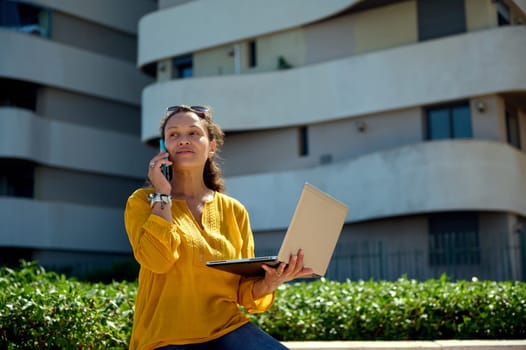 Beautiful multitasking woman, a real estate agent talking on smart mobile phone, holding laptop, standing against modern high rise buildings background. People. Online business. Remote job. Lifestyle