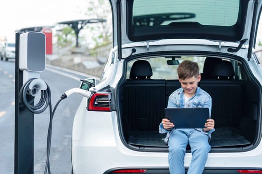 Little boy sitting on car trunk, using tablet while recharging eco-friendly car from EV charging station. EV car road trip travel as alternative vehicle using sustainable energy concept. Perpetual
