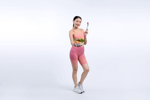 Young sporty Asian woman in sportswear holding salad bowl fill with vibrant of fruit and vegetable. Natural youthful and fit body lifestyle with balance nutrition on isolated background. Vigorous