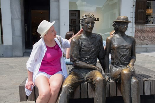 A middle-aged woman sits on a bench with two bronze statues Hendrik and Katrien, Sculpture by artist Marc Cox with the official title Listening to the carillon, Hasselt, Belgium, July 8, 2023