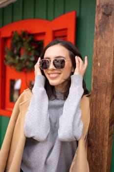 People, season and Christmas concept portrait of happy shy smiling middle age woman outdoors dressed stylish cream trench coat with Christmas festive decoration on background. Vertical photo,