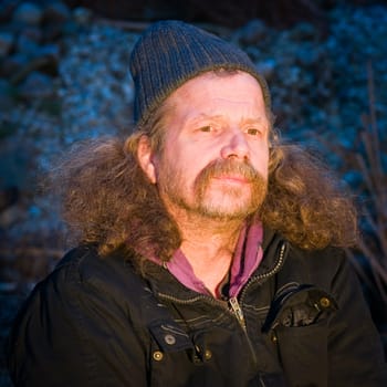 Head and shoulders portrait of an elderly man with moustache,long hair and woolly cap looking past the camera at dusk
