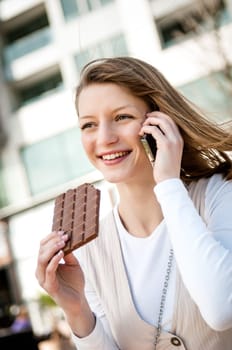 Outdoor portrait of young smiling woman eating chocolate calling with mobile phone
