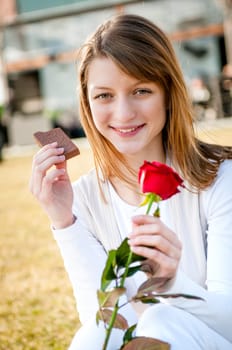 Outdoor lifestyle portrait of young woman eating chocolate with red rose