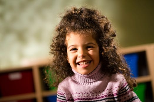 Portraits of children, happy 3 years old female with curly hair smiling and looking at camera in kindergarten