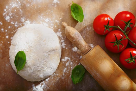 Ingredients for homemade pizza with a pile of bread dough for the base left to rise alongside fresh ripe red tomatoes and basil with a wooden rolling pin
