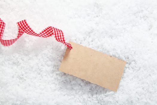 Blank paper gift tag or label with copyspace for your Christmas greeting in winter snow with a red and white checked decorative ribbon