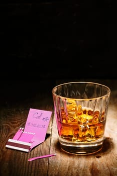 Romantic tryst on a matchbook alongside a glowing glass of whiskey on an old wooden counter in a nightclub