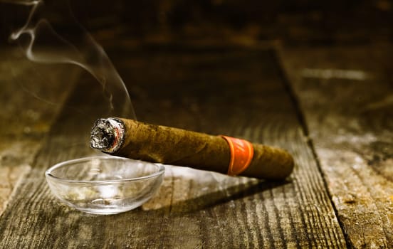 Burning handmade luxury Cuban cigar resting on an ashtray on an old wooden countertop in a nightclub or bar with copyspace