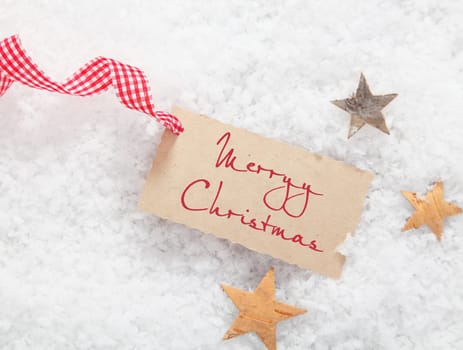 Gift tag with Merry Christmas greeting and a decorative red and white checked ribbon on a bed of winter snow with stars