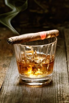 Burning Cuban cigar resting on glass of whisky on ice on an old wooden surface symbolic of masculinity
