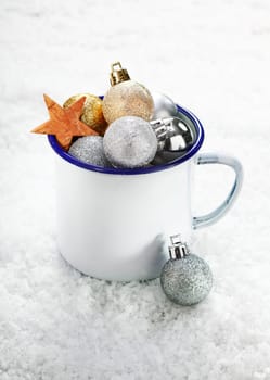 Christmas decorations in an enamelled tin mug on snow which is full to overflowing with baubles and stars
