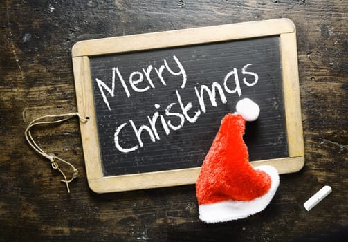 Handwritten Merry Christmas greeting on a small chalkboard with a fluffy red Santa hat on a timber background