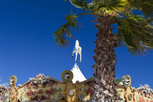 Beautiful colorful children's carousel with a peg on top stands behind a palm tree and blue sky in the marina of Gruissan in the south of France