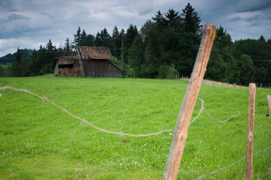 Decaying wooden hut on a green meadow in the Allgäu with barbed wire fence in Vorddergrund under cloudy skies