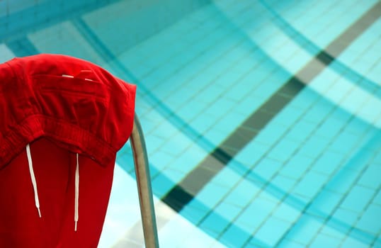 Fotografie einer roten Bermudashorts, die über das Treppengeländer eines Swimmingpools gehängt wurde. Im Hintergrund ist der Pool zu sehen.
