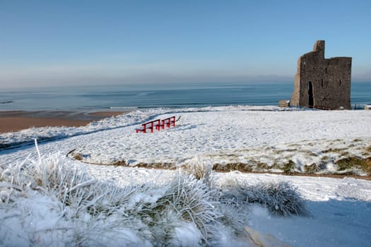 a seasonal snow covered view of atlantic ocean and ballybunion castle with red benches on a frosty snow covered winters day