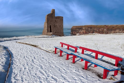 a seasonal snow covered view of atlantic ocean and ballybunion castle with red benches on a frosty snow covered winters day