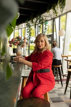 Beautiful Blonde Mature Sexagenarian Woman Drinks Coffee, Tea at Cafe or Restaurant. Modern Senior Female in her 60s wears Red Fashionable Suit, Enjoys Her Life, Time. Happy Retirement. Vertical Plane