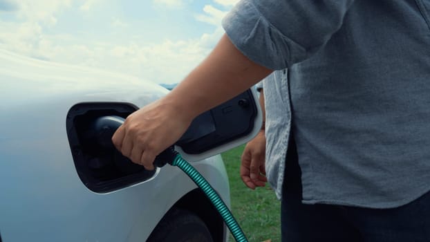 Hand insert EV charger and recharge electric car from charging station on nature and travel concept background. Technological advancement of alternative energy sustainability and EV car. Peruse