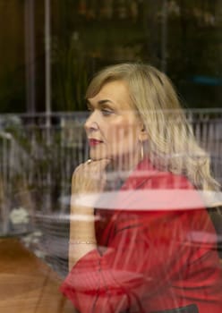 Portrait Beautiful Blonde Mature Sexagenarian Woman Sitting Behind Window Glass of Restaurant or Cafe. Stylish Modern Senior Female in her 60s Thinking About Something. Retirement. Vertical Plane.