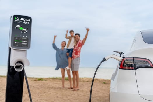 Alternative family vacation trip traveling by the beach with electric car recharging battery from EV charging station with blurred cheerful and happy family enjoying the seascape background. Perpetual