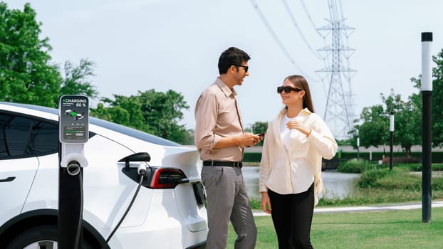 Young couple recharge EV car battery at charging station connected to power grid tower electrical industrial facility as electrical industry for eco friendly vehicle utilization. Expedient