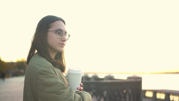 Young woman with a glass of coffee in her hand. A girl in glasses near the fence on the embankment with a hot drink in a glass. 4k