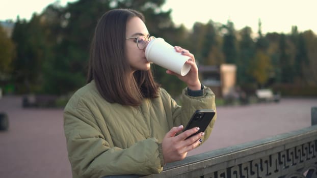 A young woman is chatting on a smartphone with a glass of coffee in her hand. A girl in glasses with a phone near the fence on the embankment with a hot drink in a glass. 4k