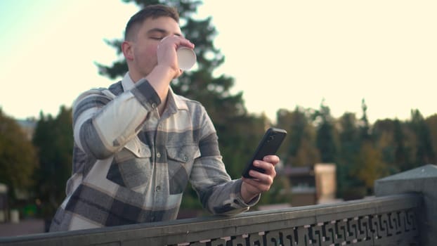 A young man is chatting on a smartphone with a glass of coffee in his hand. A man with a phone near the fence on the embankment with a hot drink in a glass. 4k