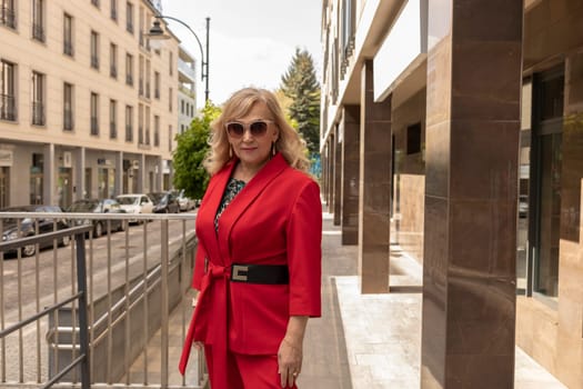 Stylish Woman In Her 60s, Long Curly Hairdo Wearing Red Jacket, Trousers, Glasses With Chain Looks Straight, Posing In City. Autumn, Summer Or Spring Fashion Personal Style. Horizontal Plane