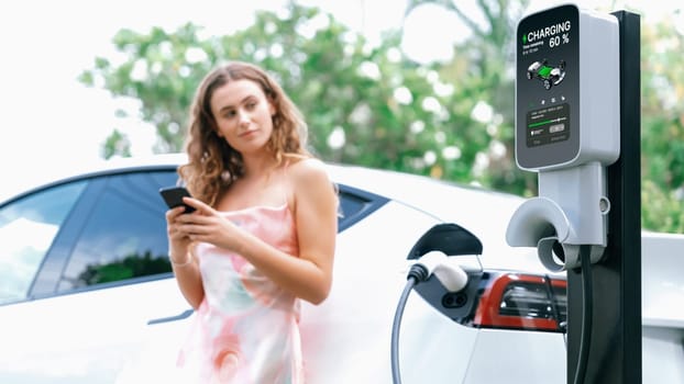 Focused charging station recharging electric vehicle on blurred background of modern woman using smartphone. EV technology utilization for tracking energy usage to optimize battery charging.Synchronos