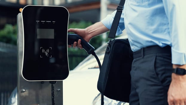 Progressive businessman install charger plug from charging station to his electric car before driving around city center. Eco friendly rechargeable car powered by sustainable and clean energy.