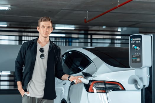 Young man travel with EV electric car to shopping center parking lot charging in downtown city showing urban sustainability lifestyle by green clean rechargeable energy of electric vehicle innards