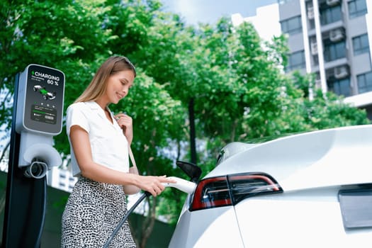 Young woman travel with EV electric car charging in green sustainable city outdoor garden in summer. Urban sustainability lifestyle by green clean rechargeable energy of electric BEV vehicle innards