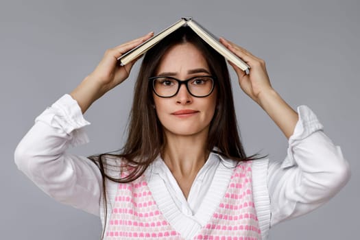 puzzled brunette woman in glasses feels sorry biting lips on grey background. oops. girl with book on head
