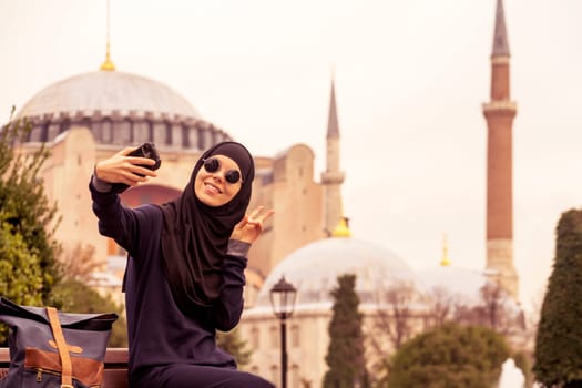 A young smiling Muslim girl in a hijab and stylish black glasses makes a selfie against the background of the Hagia Sophia mosque. Tourism in Istanbul, Turkey