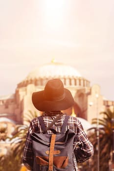 A young girl in a plaid shirt, with a stylish backpack and a hat and looks at the Hagia Sophia mosque at sunset. Istanbul, Turkey.