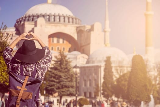 Young traveler in a hat and stylish shirt folds her hands in the shape of a heart against the background of an ancient Hagia Sophia mosque in Istanbul, Turkey. Girl tourist travels to Arab countries.