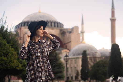 A young pretty girl in a stylish hat and a plaid shirt poses, makes a selfie on the phone next to the Hagia Sophia mosque. Tourism in Istanbul, Turkey.