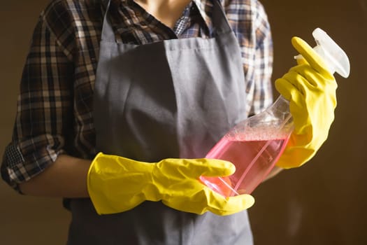 Young female hands in yellow rubber gloves hold a new detergent for cleaning the house and wiping down surfaces. A housekeeper in a plaid shirt and gray apron holds a rag and plans to disinfect.