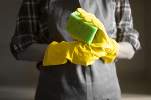 Young female hands in yellow rubber gloves hold a new green sponge for cleaning house and wiping surfaces. A female housekeeper in a plaid shirt and a gray apron holds a rag and plans to disinfect.