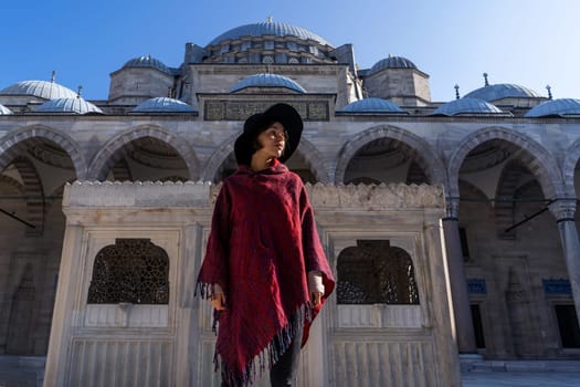 A young girl traveler in a stylish red poncho and a hat stands against the background of a blue mosque in Istanbul, Turkey. Backpacker travels to Arab countries