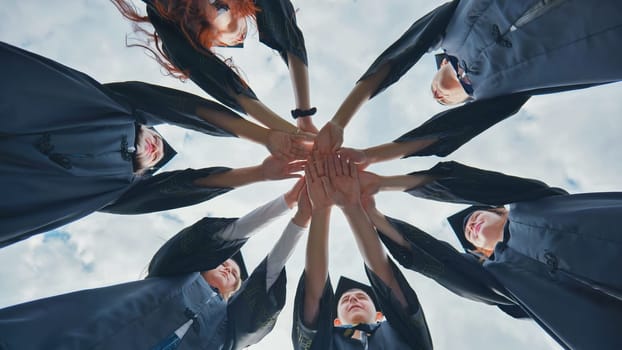 Team of college or university students celebrating graduation. Group of happy successful graduates in academic hats and robes standing in circle and putting their hands together