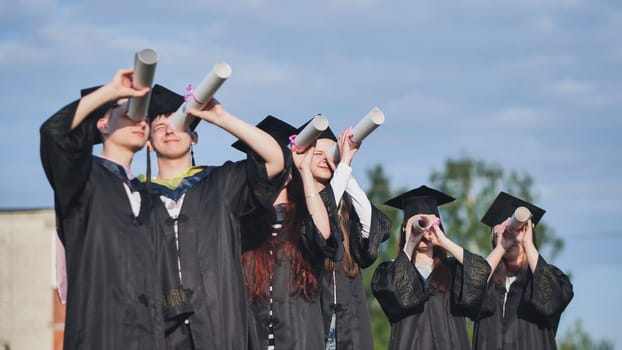 Cheerful graduates on a sunny day look through diplomas like a telescope