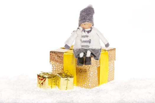 Little doll sitting on Christmas gift with small golden packages in the snow against white background
